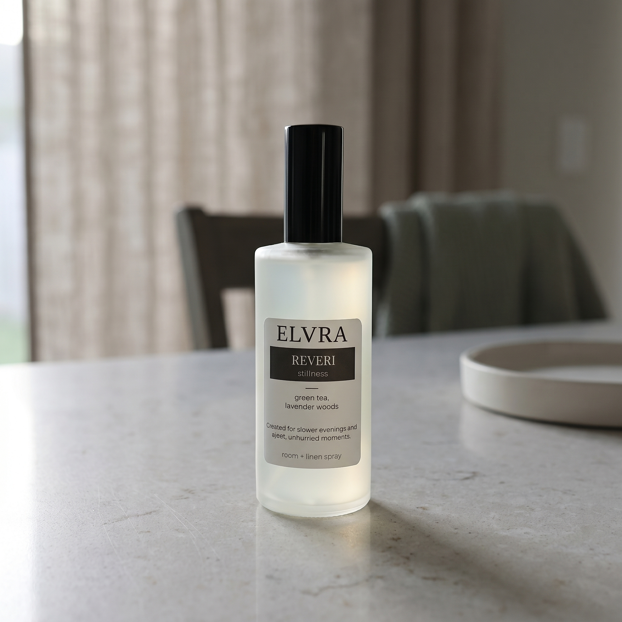 Frosted Elvra Reveri atmosphere mist bottle centered on pale stone counter with soft dusk light, linen curtains, and muted sage accents.