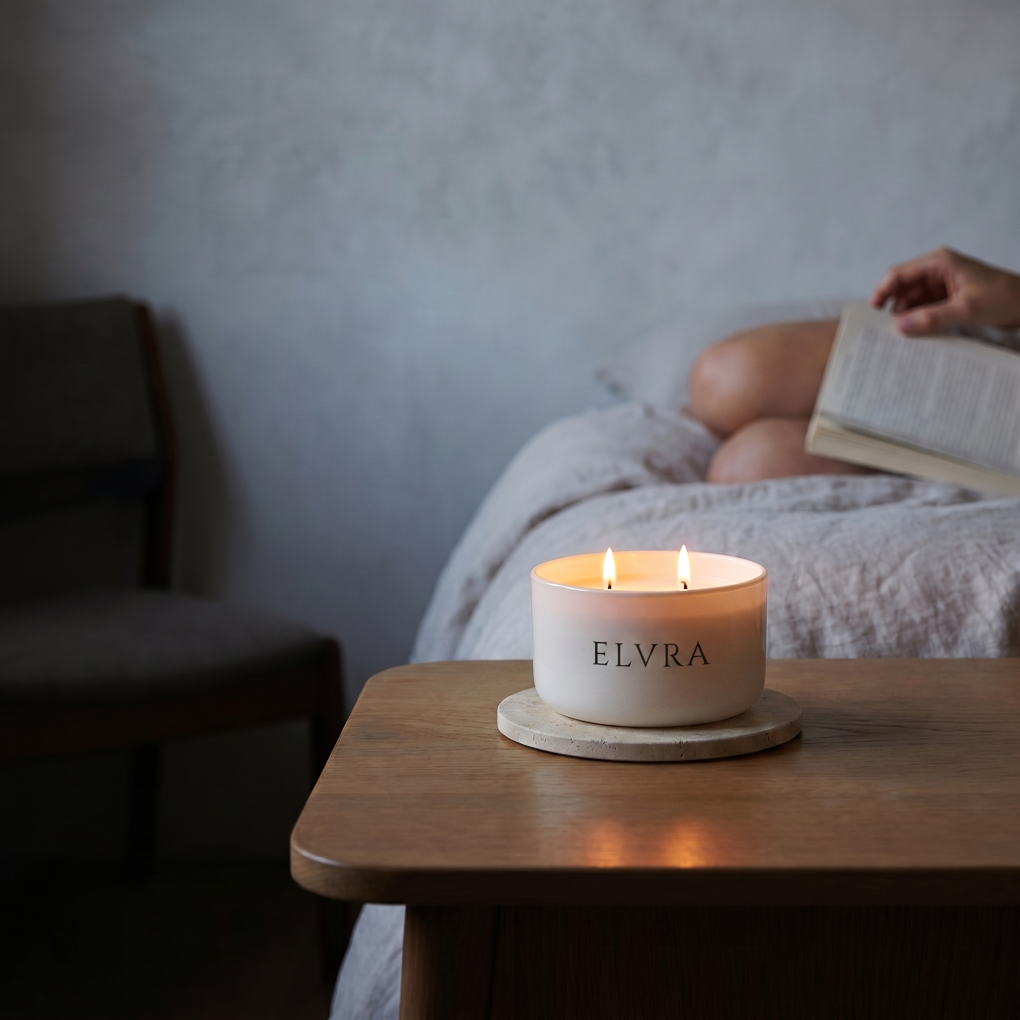 Candle with 'ELVRA' branding on a wooden table, person reading a book in the background