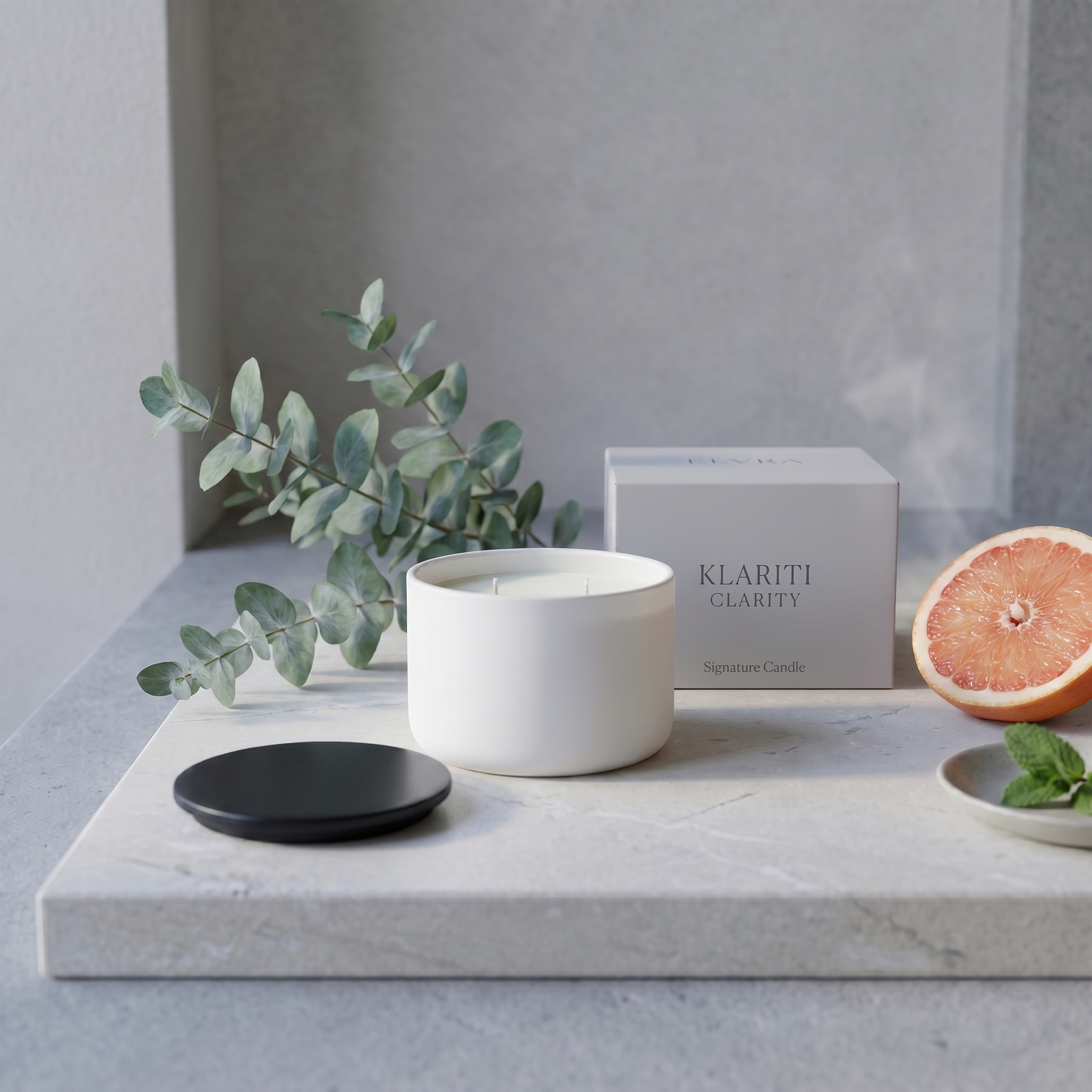 A serene luxury candle still life with a white KLARITI™ candle centered on a pale stone surface, framed by soft green botanicals, a grapefruit slice, and a black lid in gentle daylight.