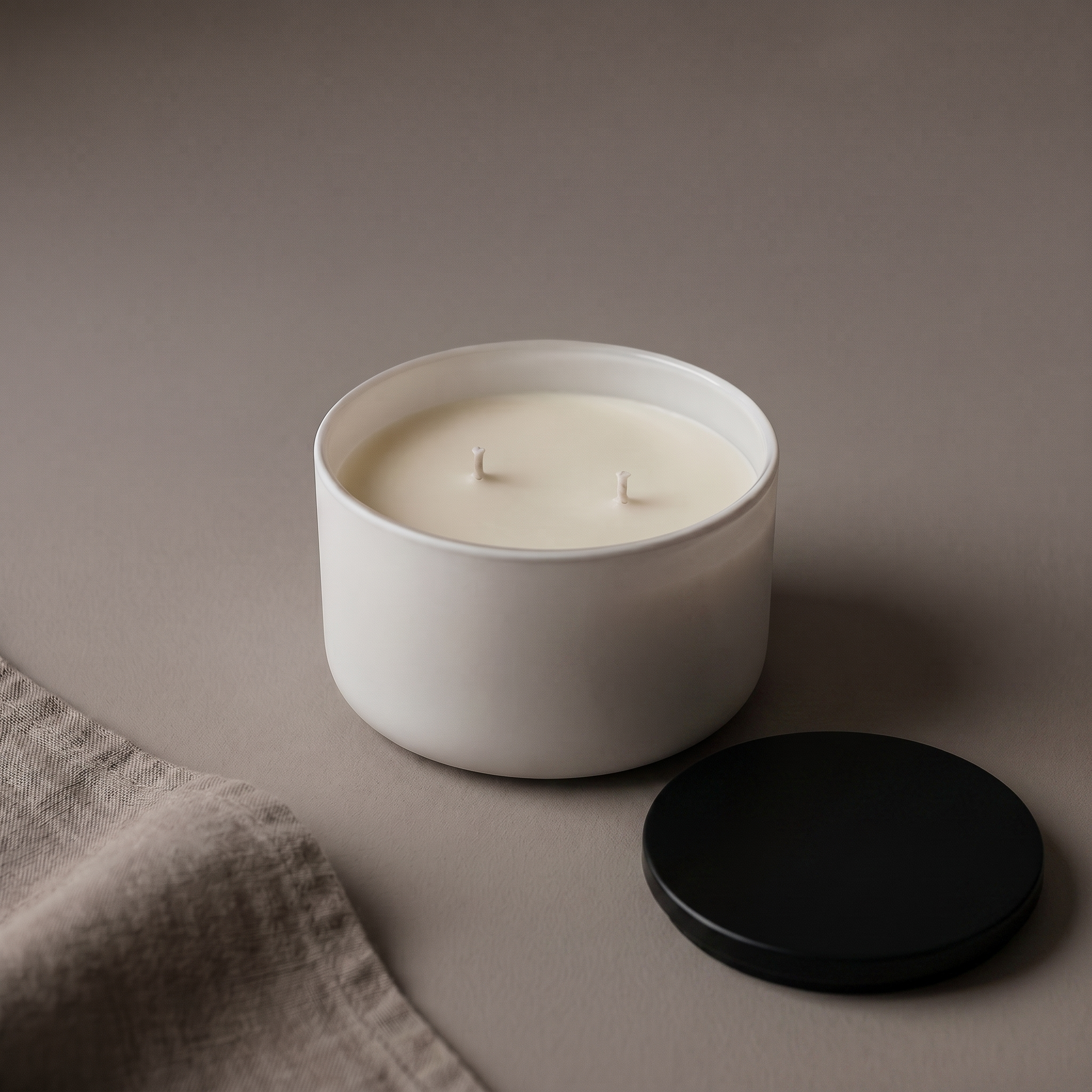 Minimal white candle on a warm beige surface with a black lid placed nearby, creating a quiet, premium still life with soft studio shadows.